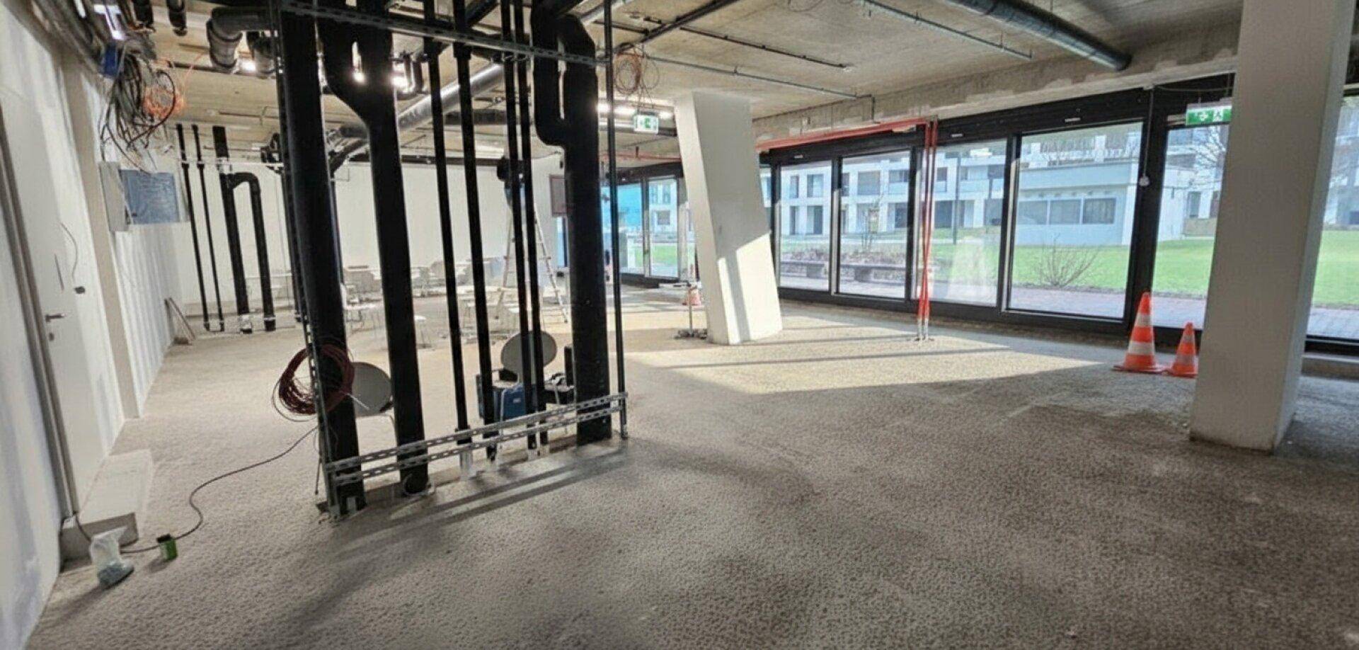 Bürofläche im Bau mit sichtbaren Leitungen, hohen Decken und großen Fenstern, die viel Licht hereinlassen.