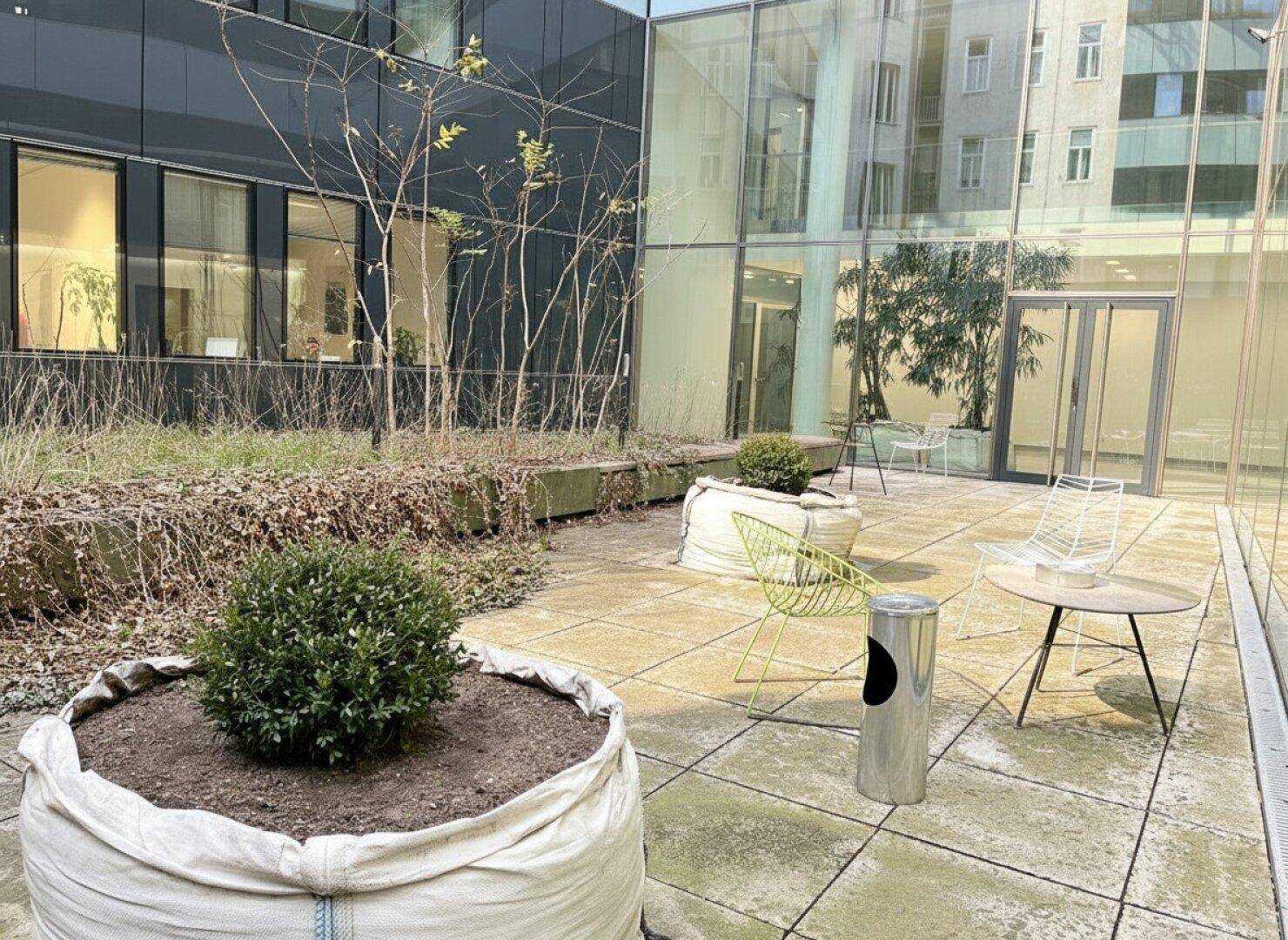 Ruhiger Innenhof mit gepflastertem Bereich, kleinen Pflanzen und modernen Sitzgelegenheiten vor einer Glasfassade.
