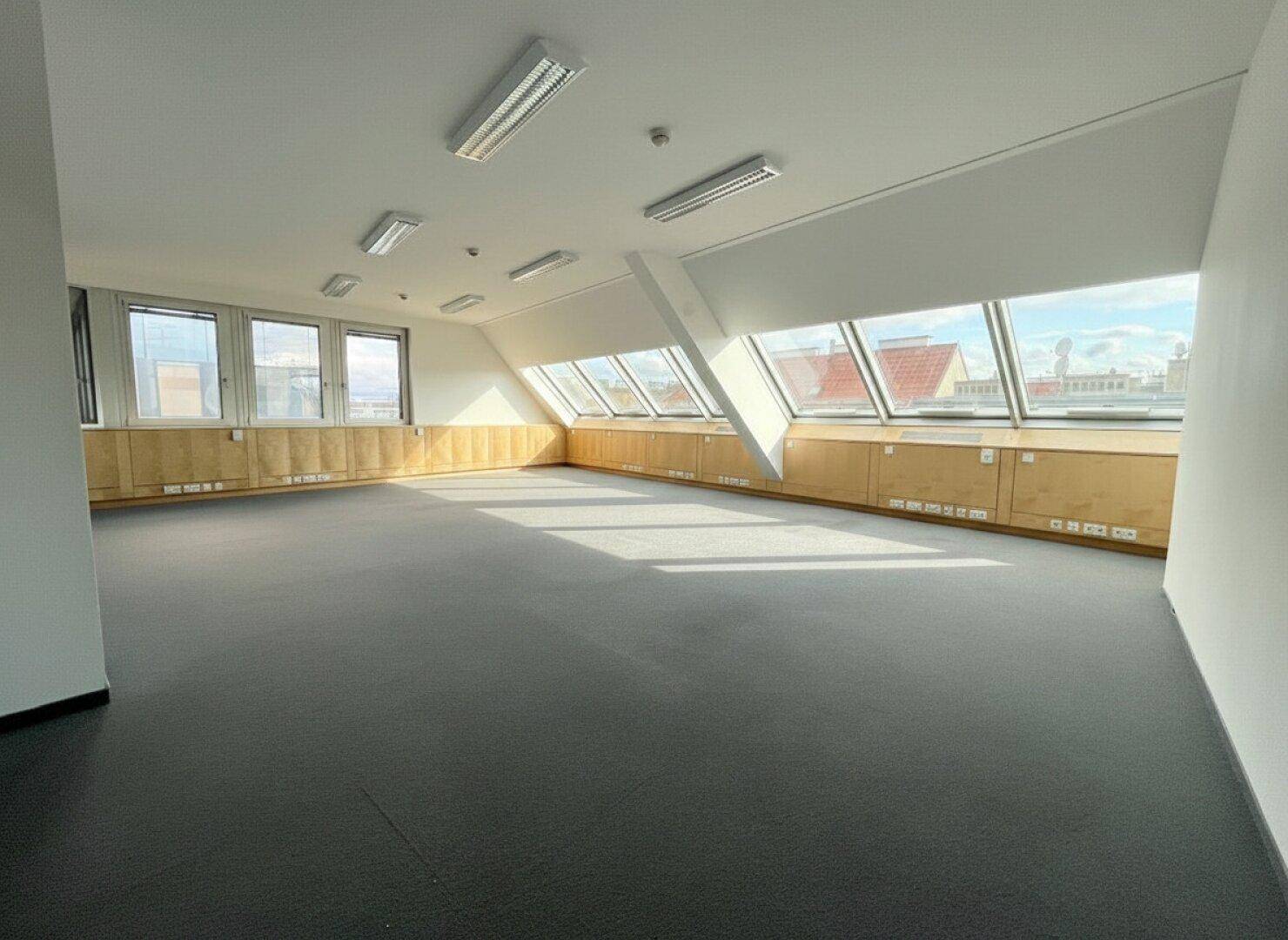 Geräumiges Büro im Dachgeschoss mit Schrägfenstern und Teppichboden, gut beleuchtet durch natürliches Licht.