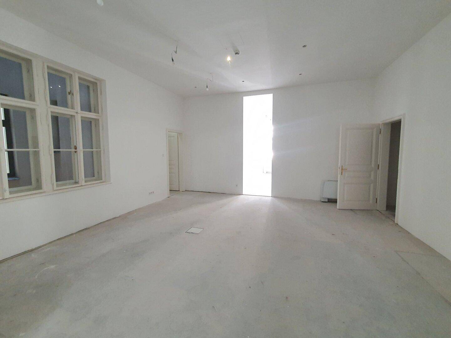Geräumiger, heller Büroraum mit einem großen Fenster und einer Tür, durchflutet von natürlichem Licht.