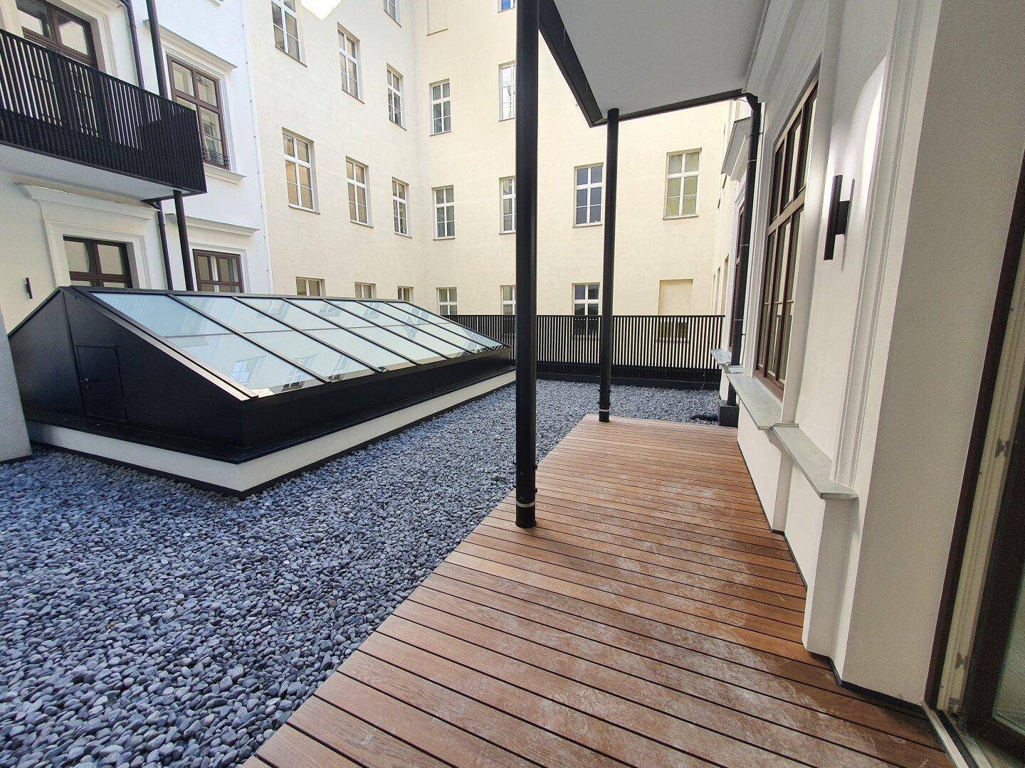 Außenbereich mit Holzterrasse, Kiesfläche und großem Oberlicht, umgeben von Gebäudefassaden.
