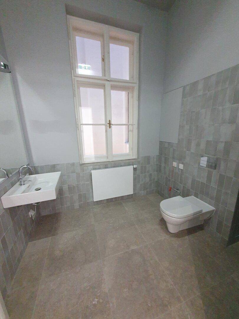 Modernes Badezimmer mit grauen Fliesen, wandhängendem WC, Waschbecken und Fenster.