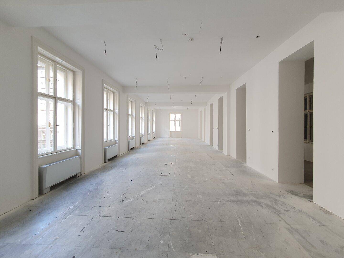 Weitläufiger, heller Büroraum mit zahlreichen Fenstern und einem schlichten Betonboden.