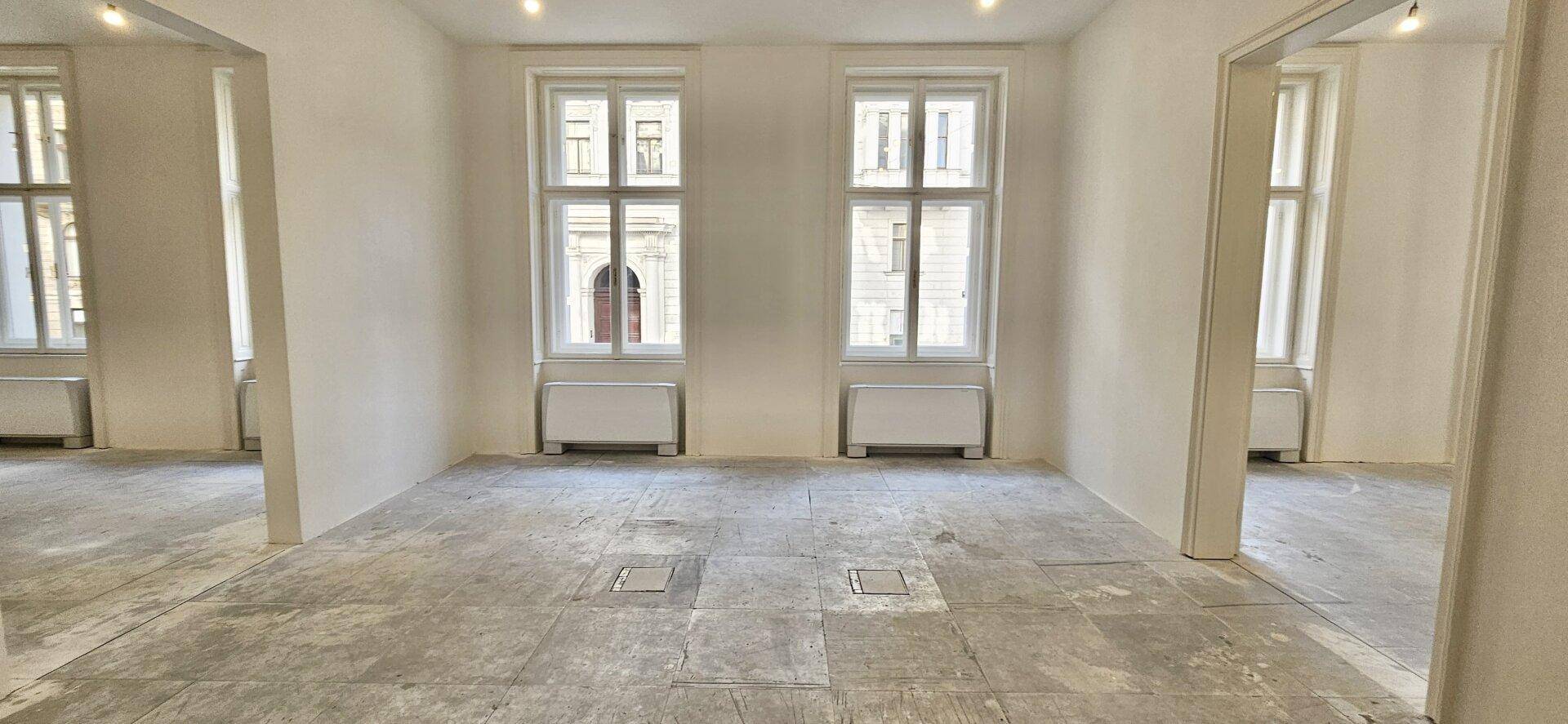 Großer Büroraum mit zwei Fenstern und Durchgängen zu weiteren Räumen, im Rohbauzustand mit Betonboden.