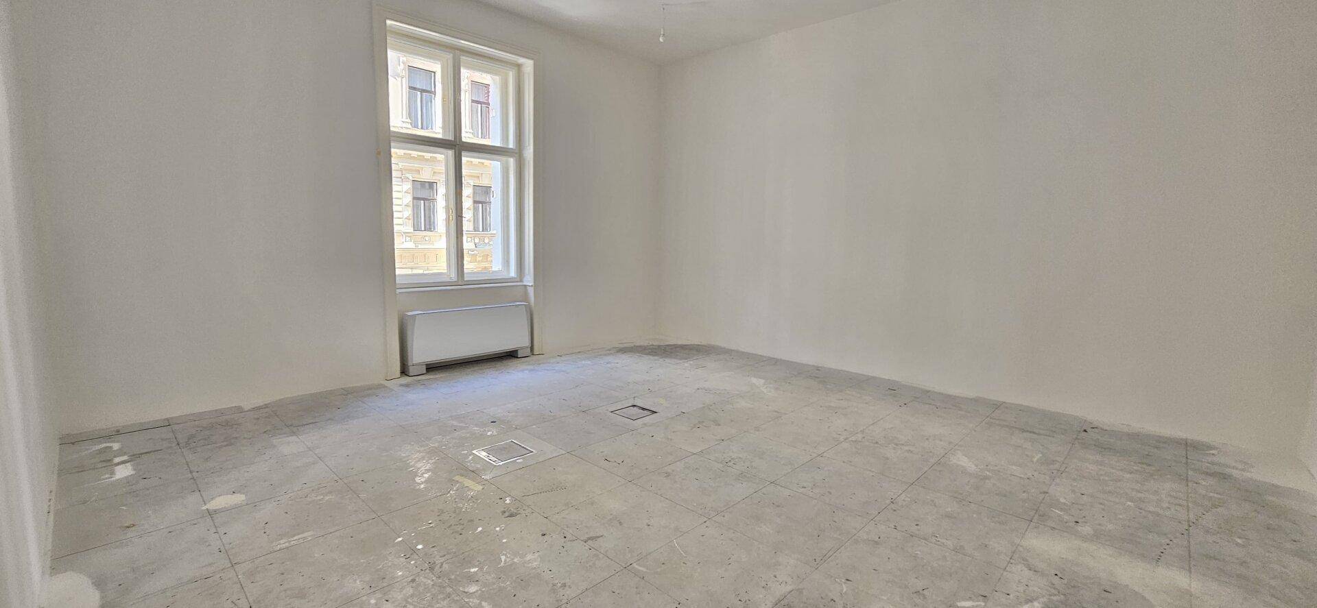 Einzelner Büroraum mit einem großen Fenster, das viel natürliches Licht spendet, im Rohbauzustand mit Betonboden.
