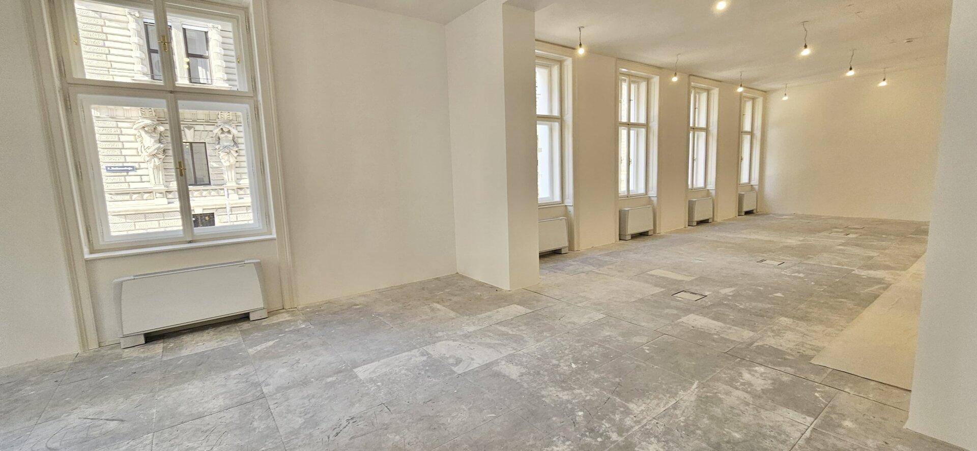 Langer Büroraum im Rohbau mit mehreren Fenstern und Betonboden, der auf den Innenausbau wartet.