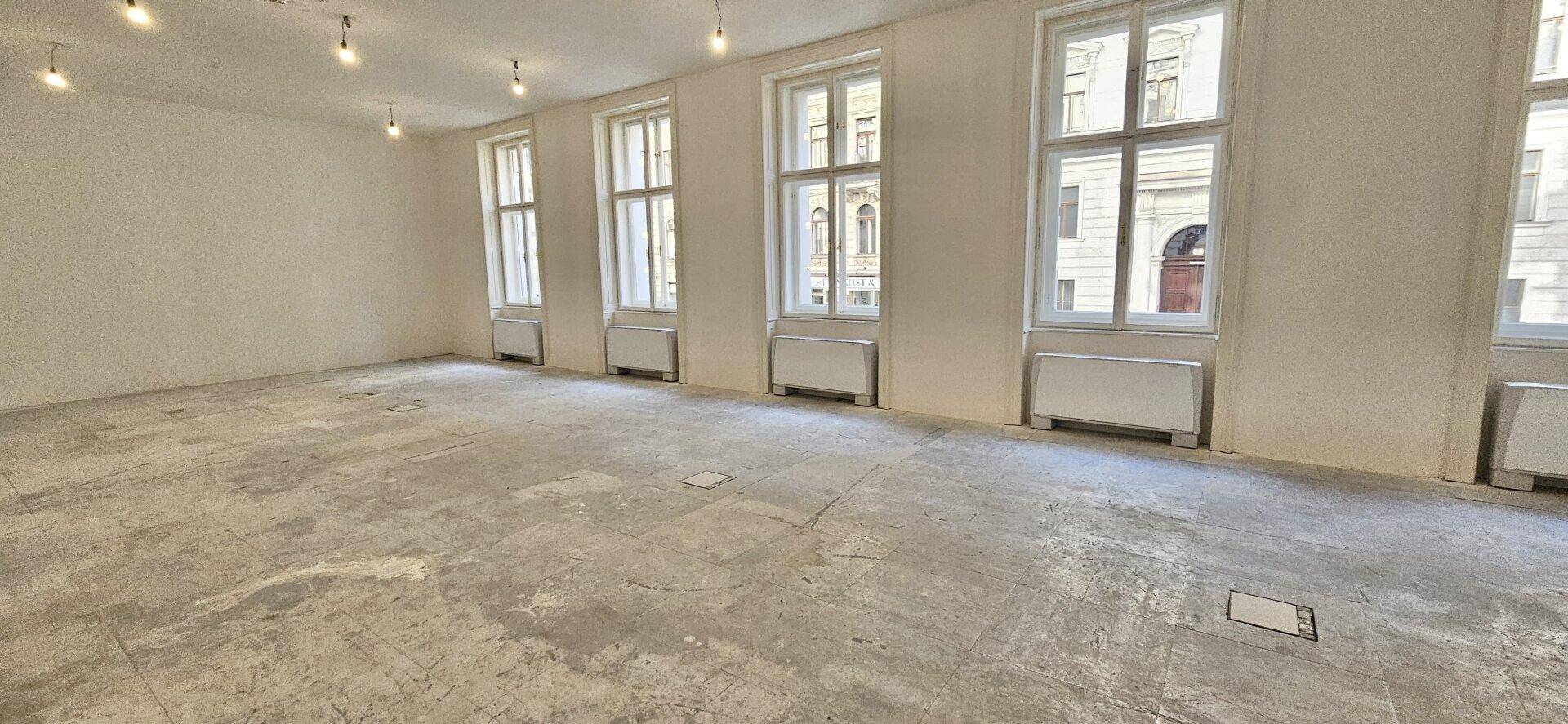 Langer Büroraum im Rohbau mit mehreren Fenstern und Betonboden, der auf den Innenausbau wartet.