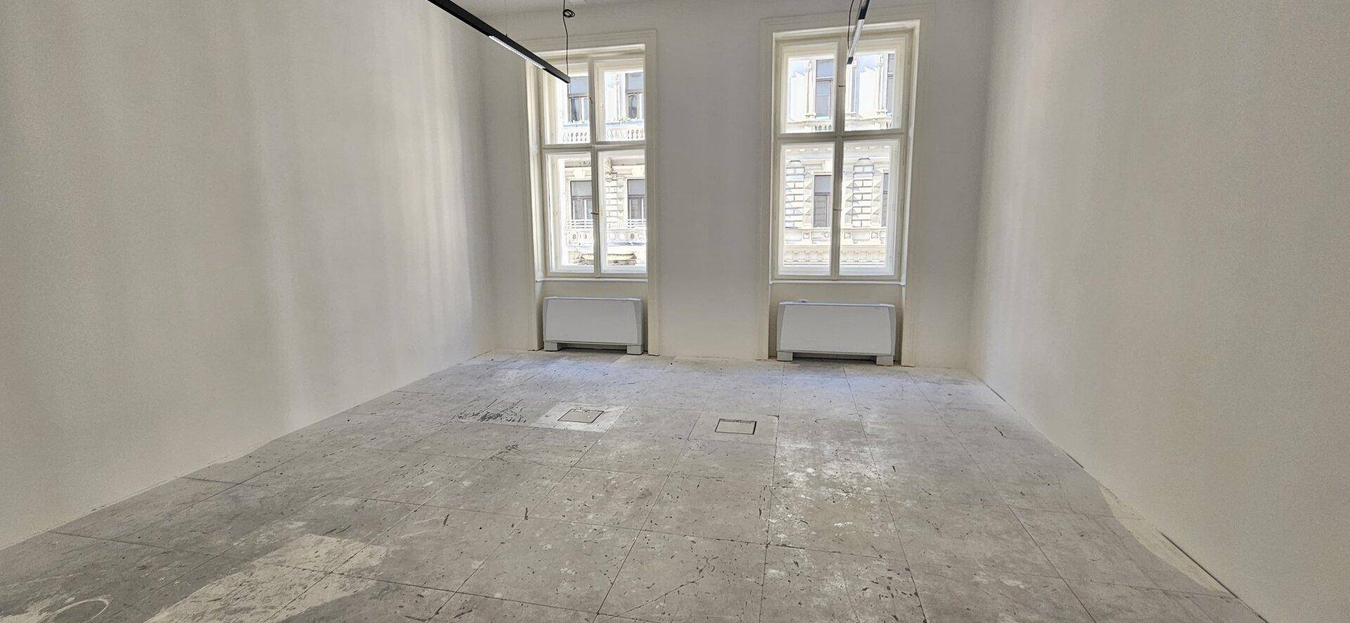 Heller Büroraum im Rohbau mit zwei großen Fenstern und Betonboden, bereit für die Gestaltung.