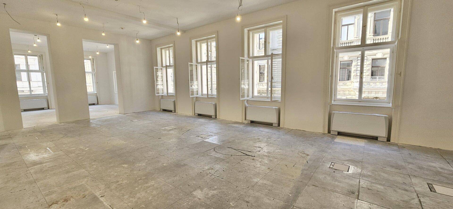 Großer Büroraum mit mehreren Fenstern und Durchgängen, die eine offene und helle Arbeitsumgebung schaffen.