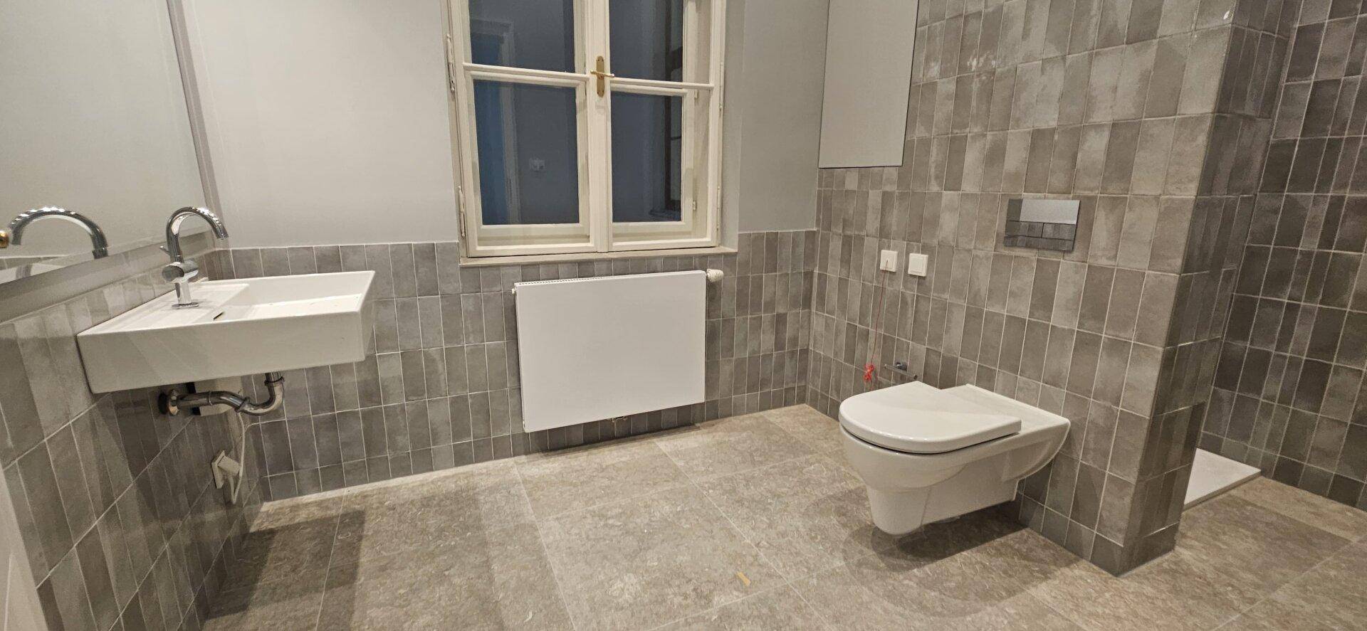 Modernes Badezimmer mit grauen Fliesen, wandhängendem WC und Waschbecken, sowie einem Fenster.