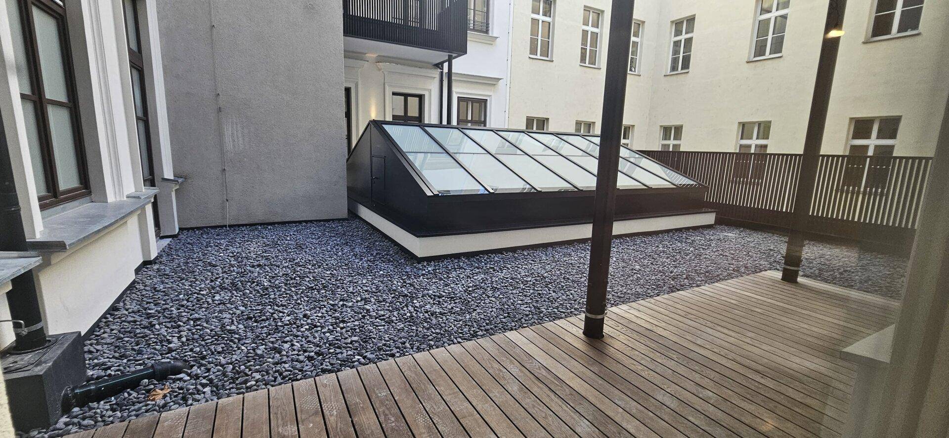 Außenbereich mit Holzdielen und Kies, sowie einem Glasdach, das Licht in den darunterliegenden Raum bringt.