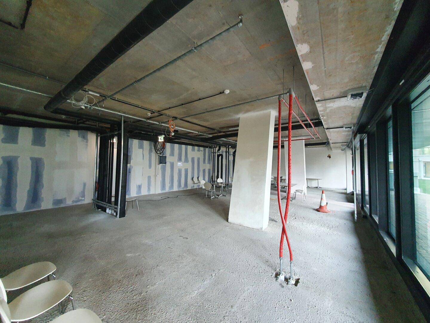 Unfertiger Büroraum mit Betonboden, sichtbaren Installationen und großen Fenstern, die viel Licht hereinlassen.