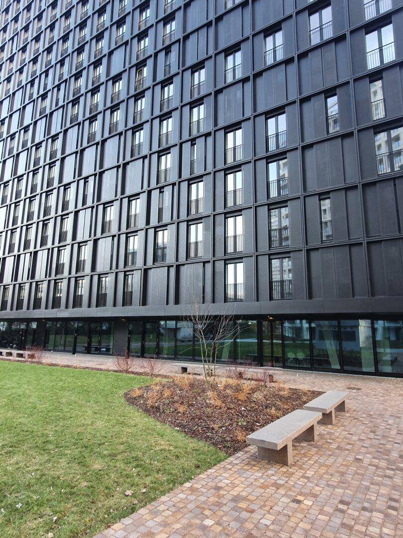 Moderne Gebäudefassade mit großen Fenstern im Erdgeschoss, umgeben von einer Grünfläche und einem Steinweg.