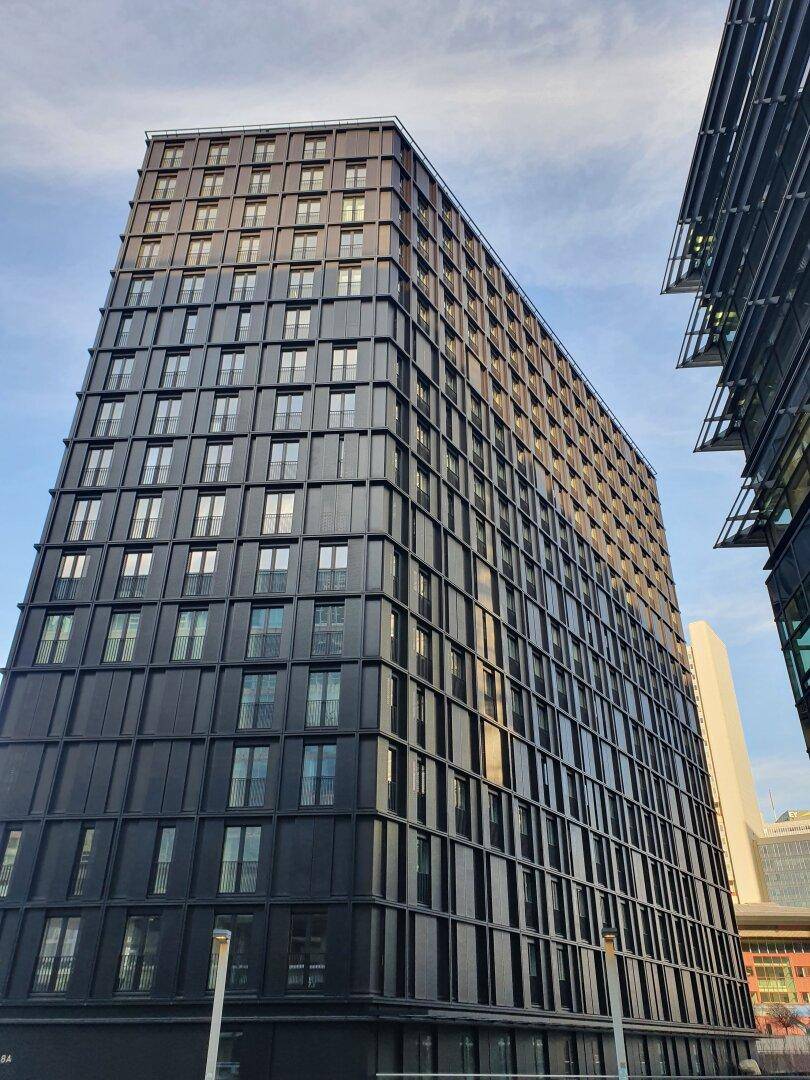 Hohes, modernes Gebäude mit dunkler Fassade und zahlreichen Fenstern unter blauem Himmel.