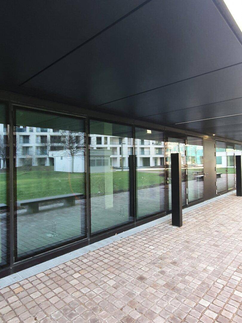 Moderne Glasfassade eines Gebäudes mit großen Fenstern und gepflastertem Vorplatz.