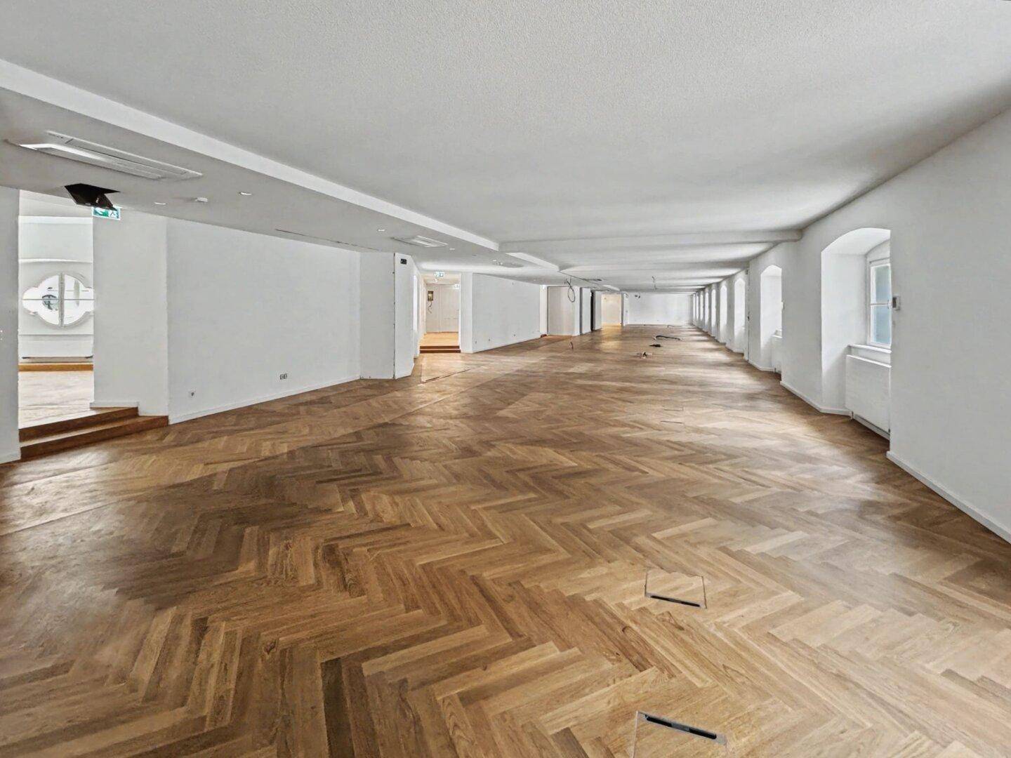 Langer, offener Bürobereich mit Fischgrätparkett und vielen Fenstern, ideal für flexible Arbeitsplatzgestaltung.