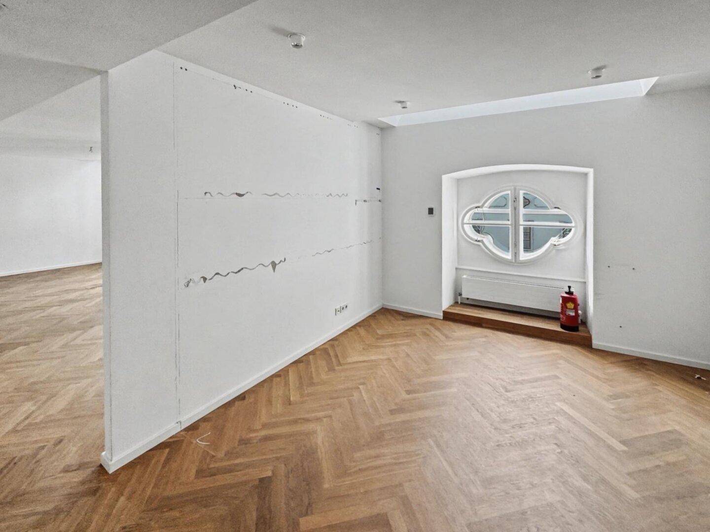 Büroraum mit Fischgrätparkett, weiß gestrichenen Wänden und einem dekorativen Rundbogenfenster, mit Feuerlöscher.