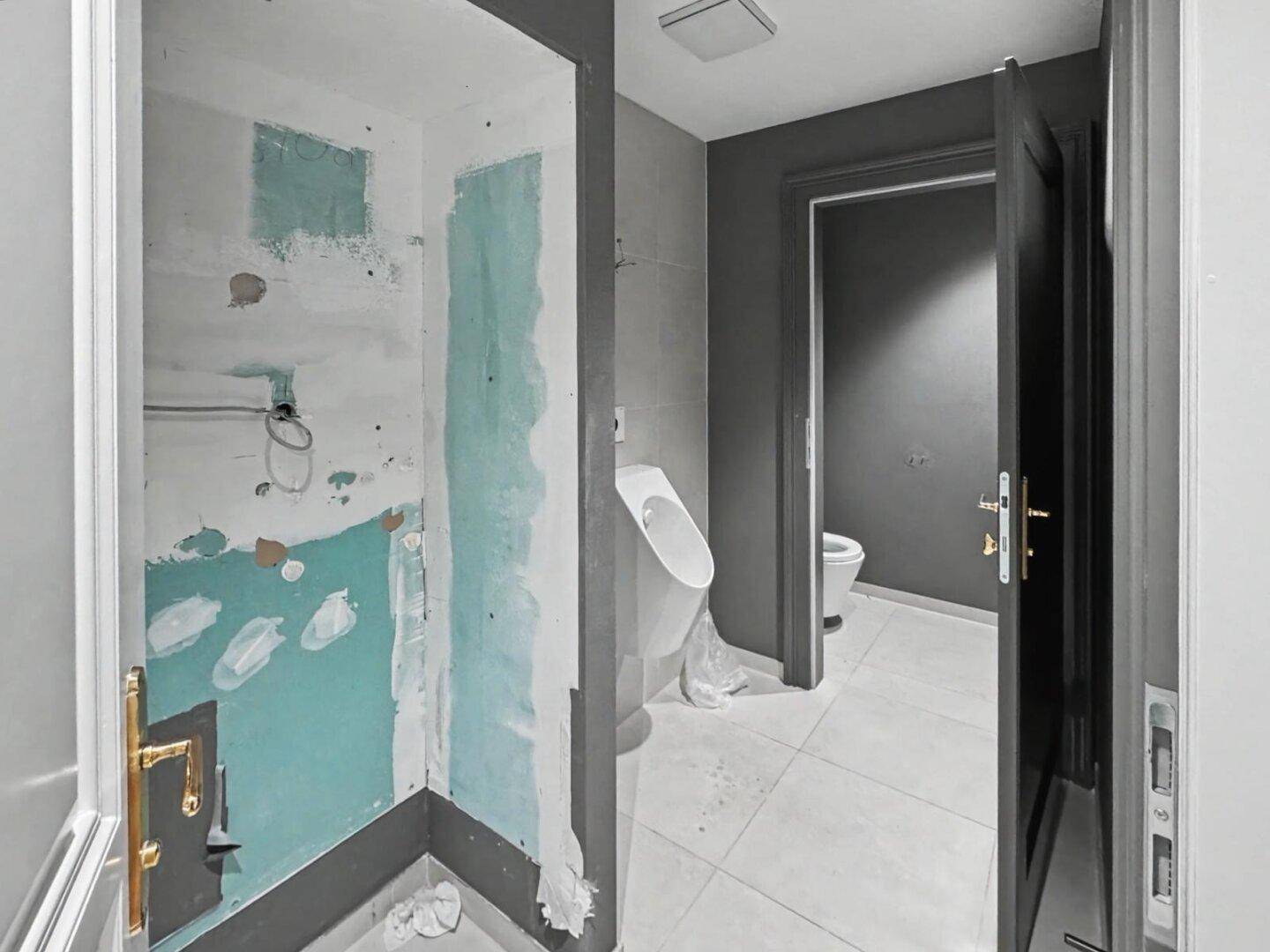 Modernes Badezimmer mit dunklen Fliesen, Urinal und Toilette, teilweise noch im Bau.