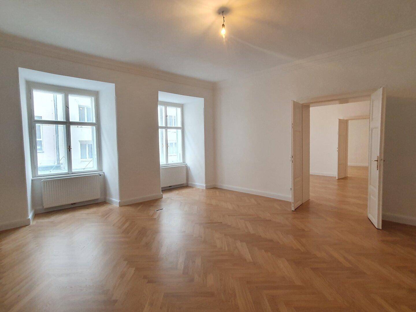 Heller Wohnraum mit klassischem Fischgrätparkett und zwei hohen Fenstern, die den Raum erhellen.