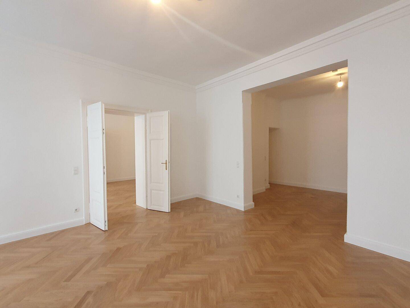 Geräumiger Wohnbereich mit Fischgrätparkett und einem breiten Durchgang zu einem weiteren Zimmer.