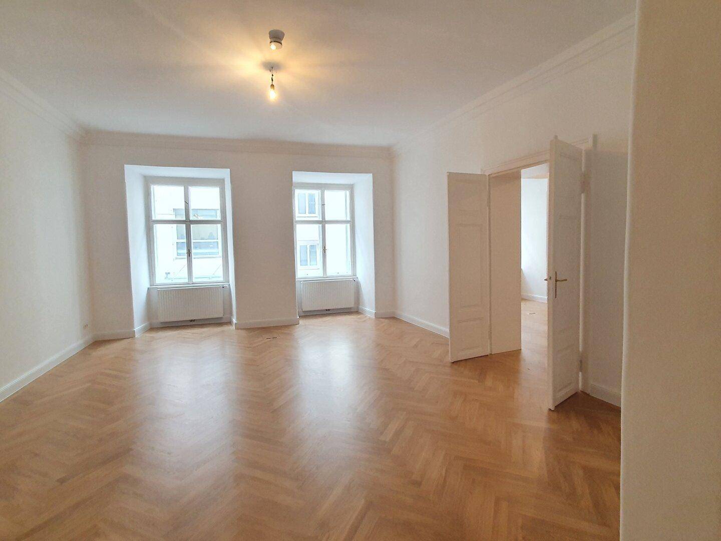 Heller Raum mit Fischgrätparkett und zwei Fenstern, die den Blick nach draußen freigeben.