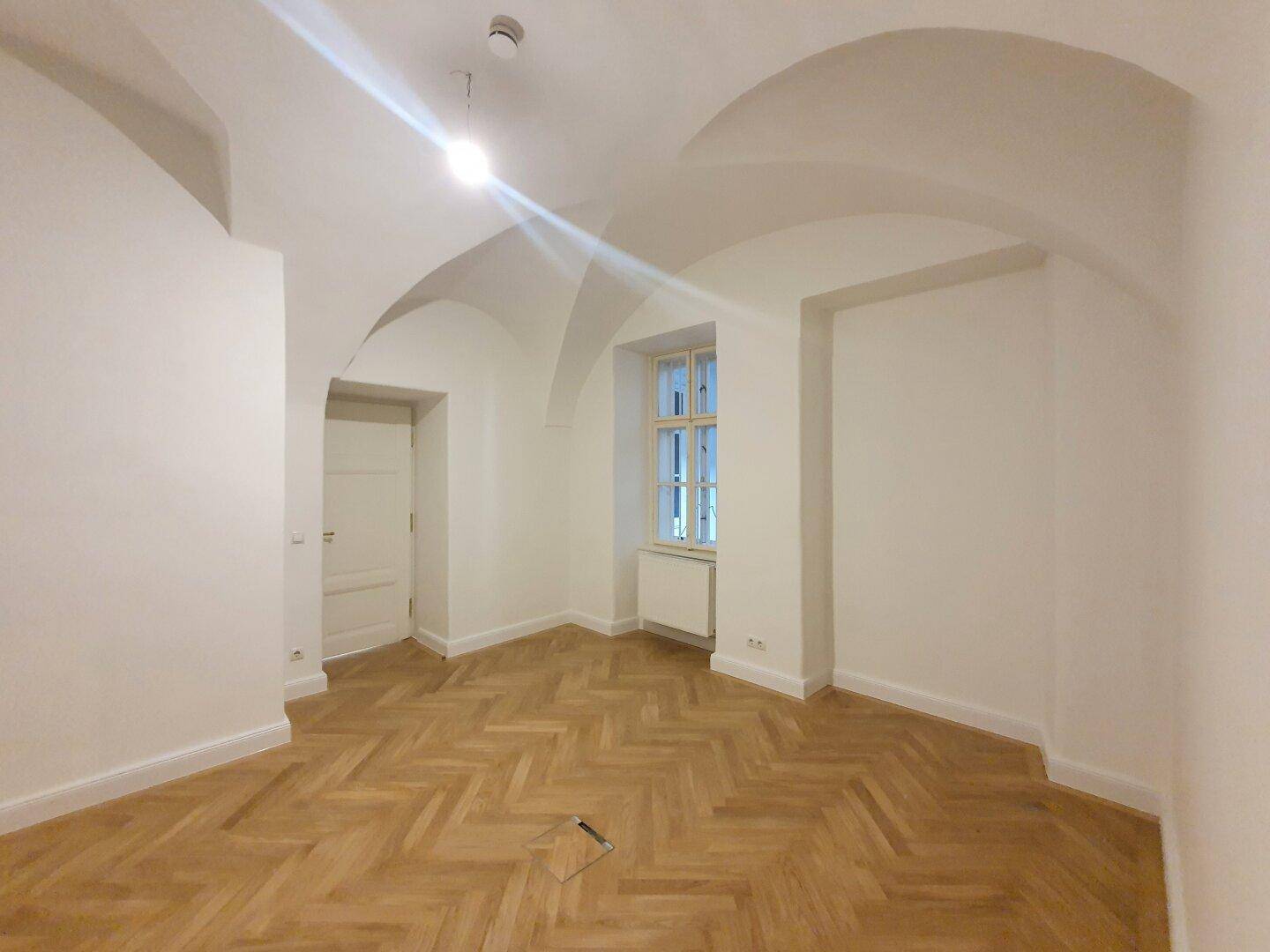 Zimmer mit beeindruckendem Gewölbedecke, Fischgrätparkett und einem Fenster, das den Raum erhellt.