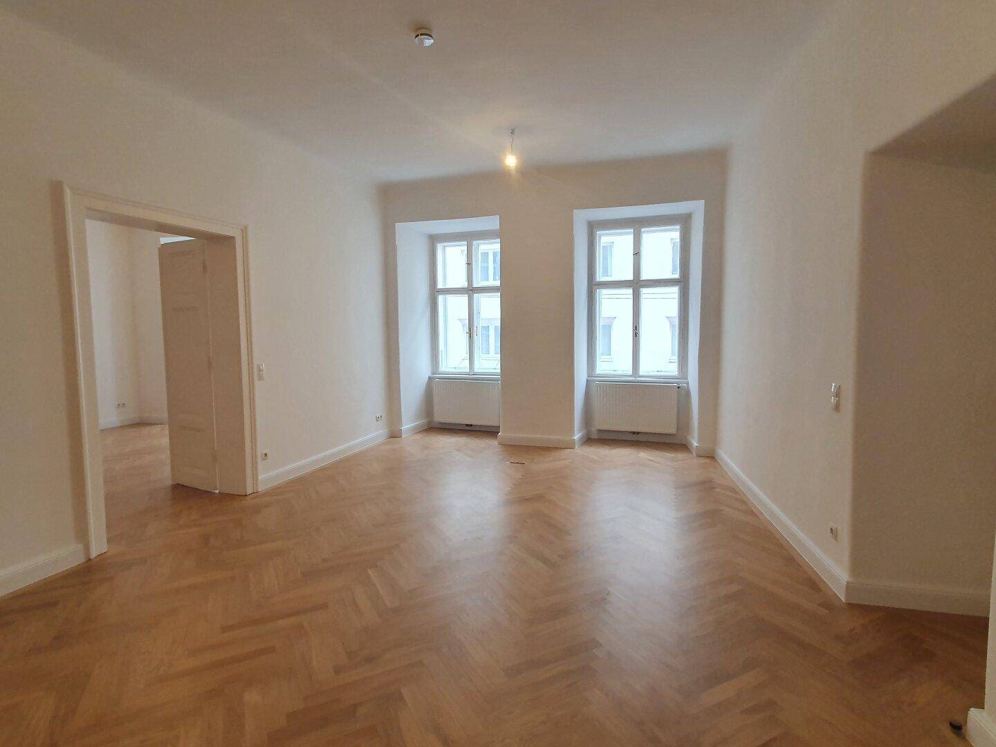 Geräumiger Raum mit Fischgrätparkett und zwei hohen Fenstern, die den Raum mit Licht durchfluten.