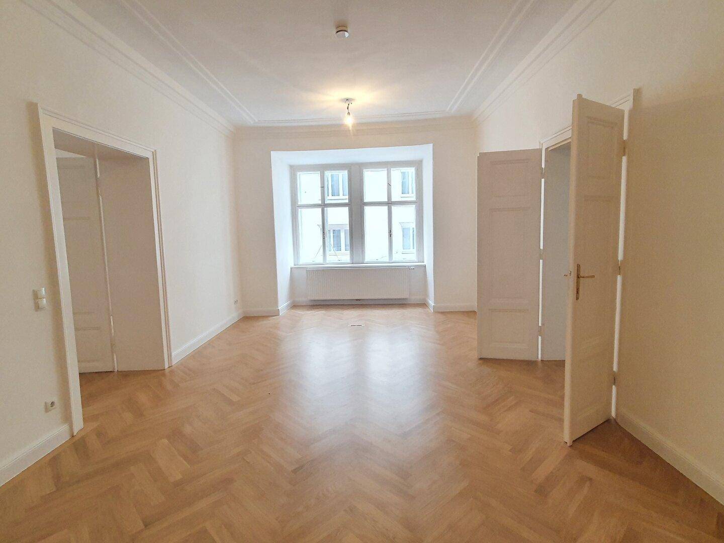 Heller Wohnraum mit Fischgrätparkett, hohen Decken und großen Fenstern, die viel natürliches Licht hereinlassen.