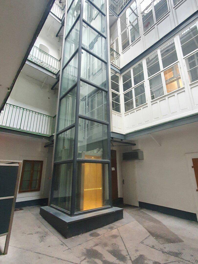 Moderner Glasaufzug im Innenhof eines Altbaus, umgeben von Fenstern und Balkonen.