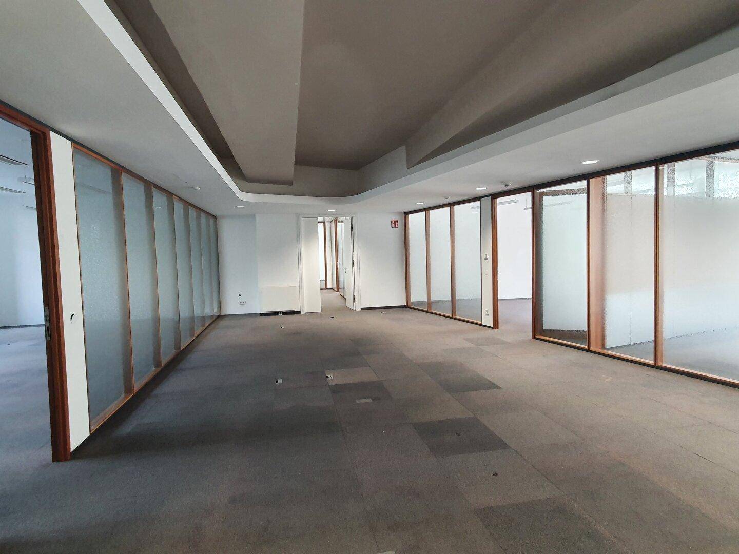 Großes Büro mit Teppichboden, Glaswänden und Zugang zu weiteren Räumen.