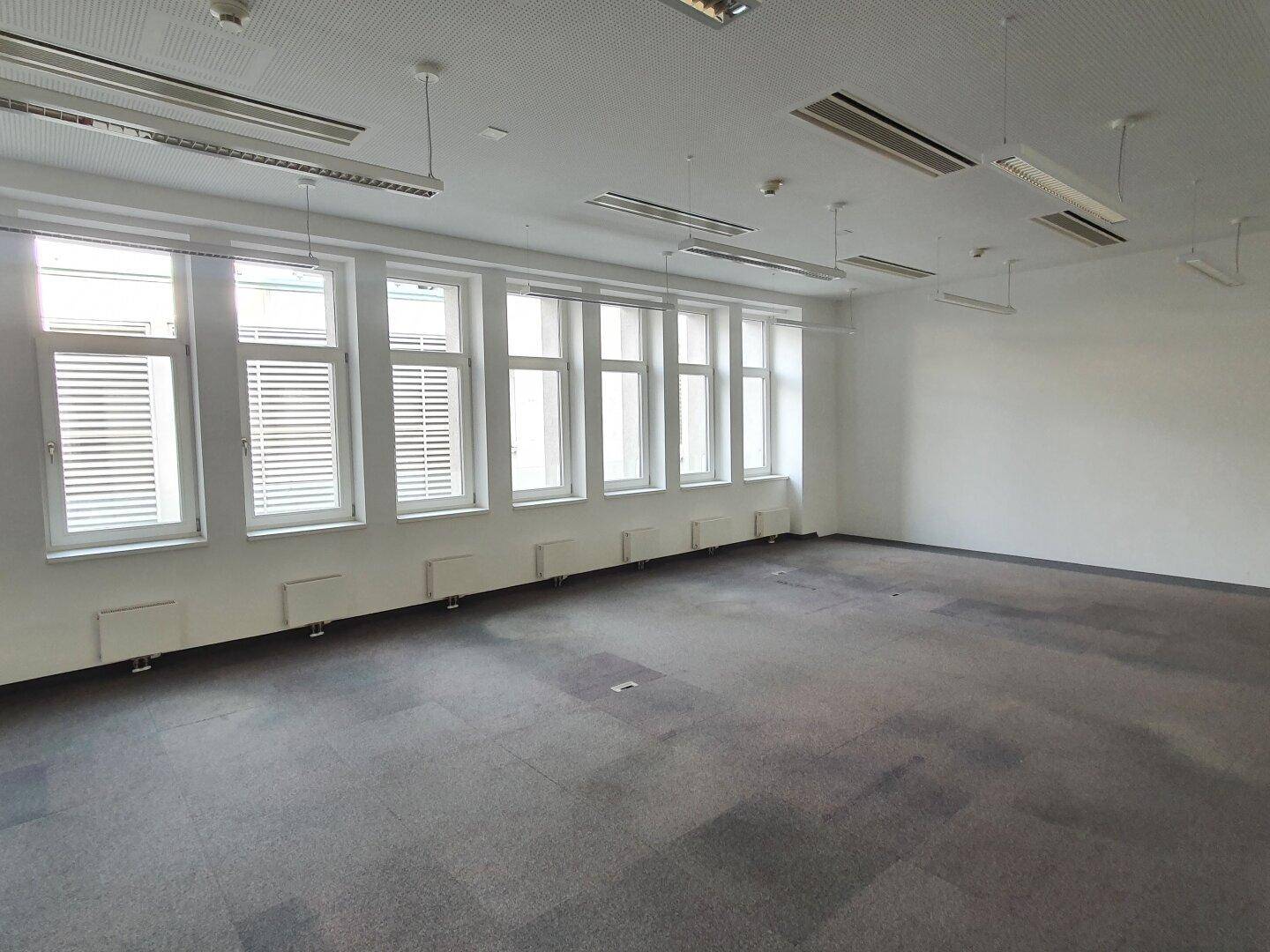 Helles Büro mit vielen Fenstern, Teppichboden und Deckenbeleuchtung.