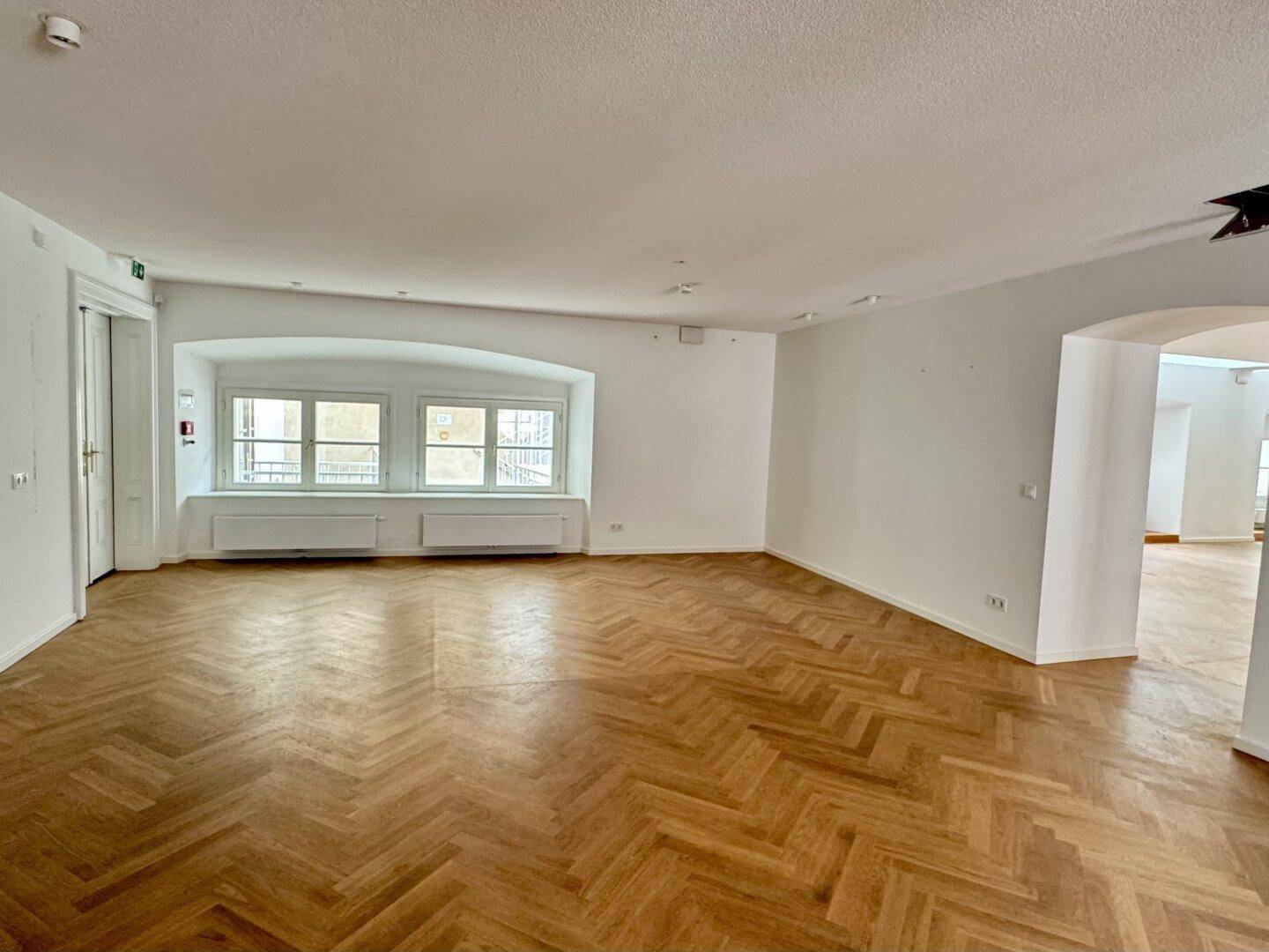 Heller Büroraum mit Fischgrätparkett und zwei großen Fenstern mit Blick nach draußen.