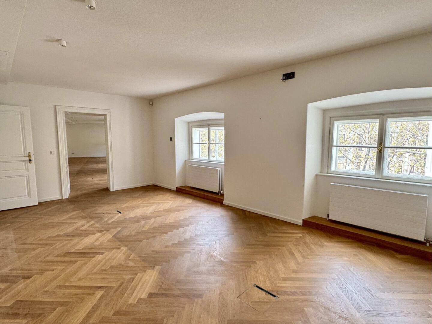 Heller Büroraum mit Fischgrätparkett, hohen Decken und Blick auf Bäume.