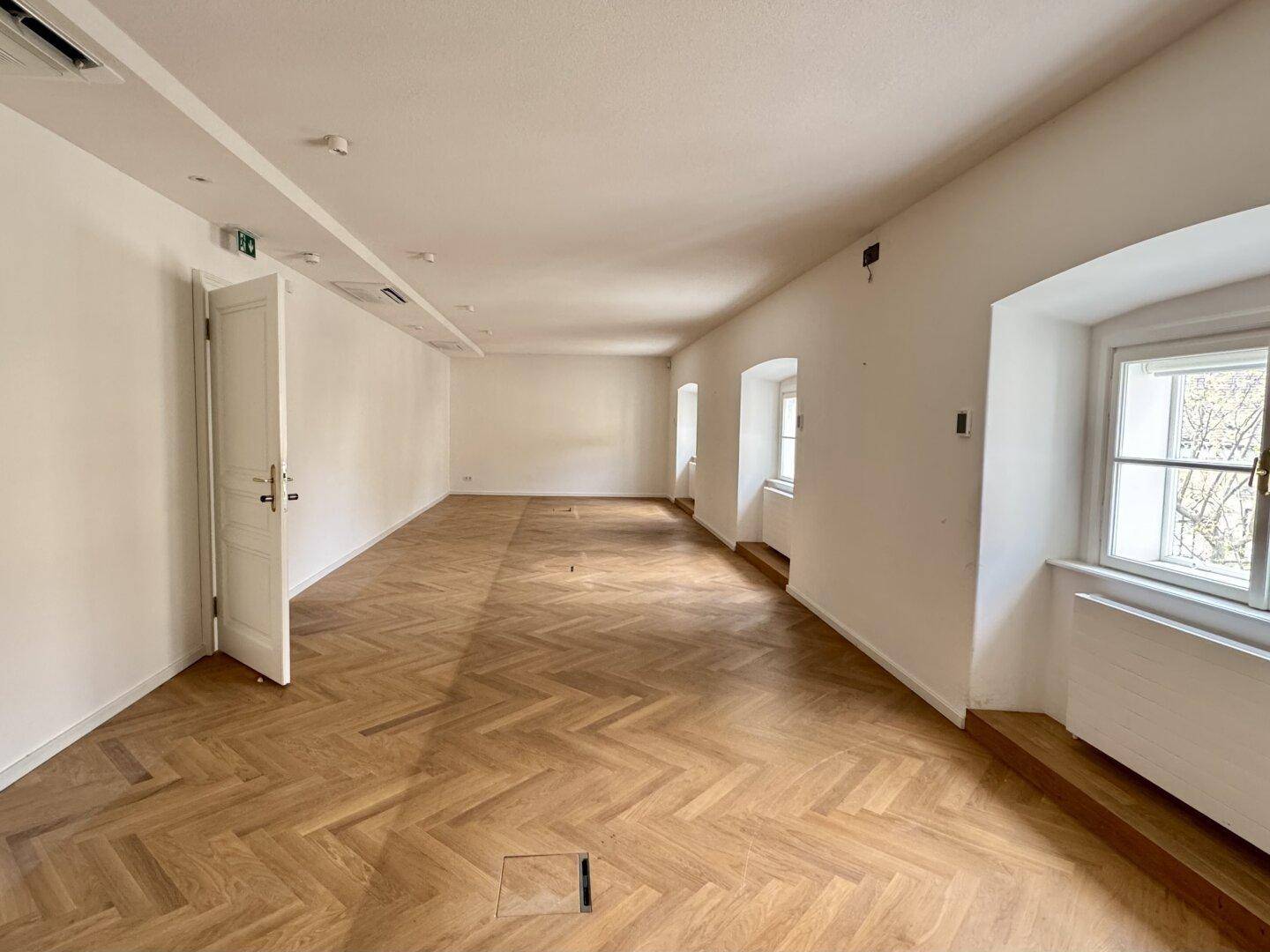 Langer, heller Büroraum mit Fischgrätparkett und mehreren Fenstern entlang der Wand.