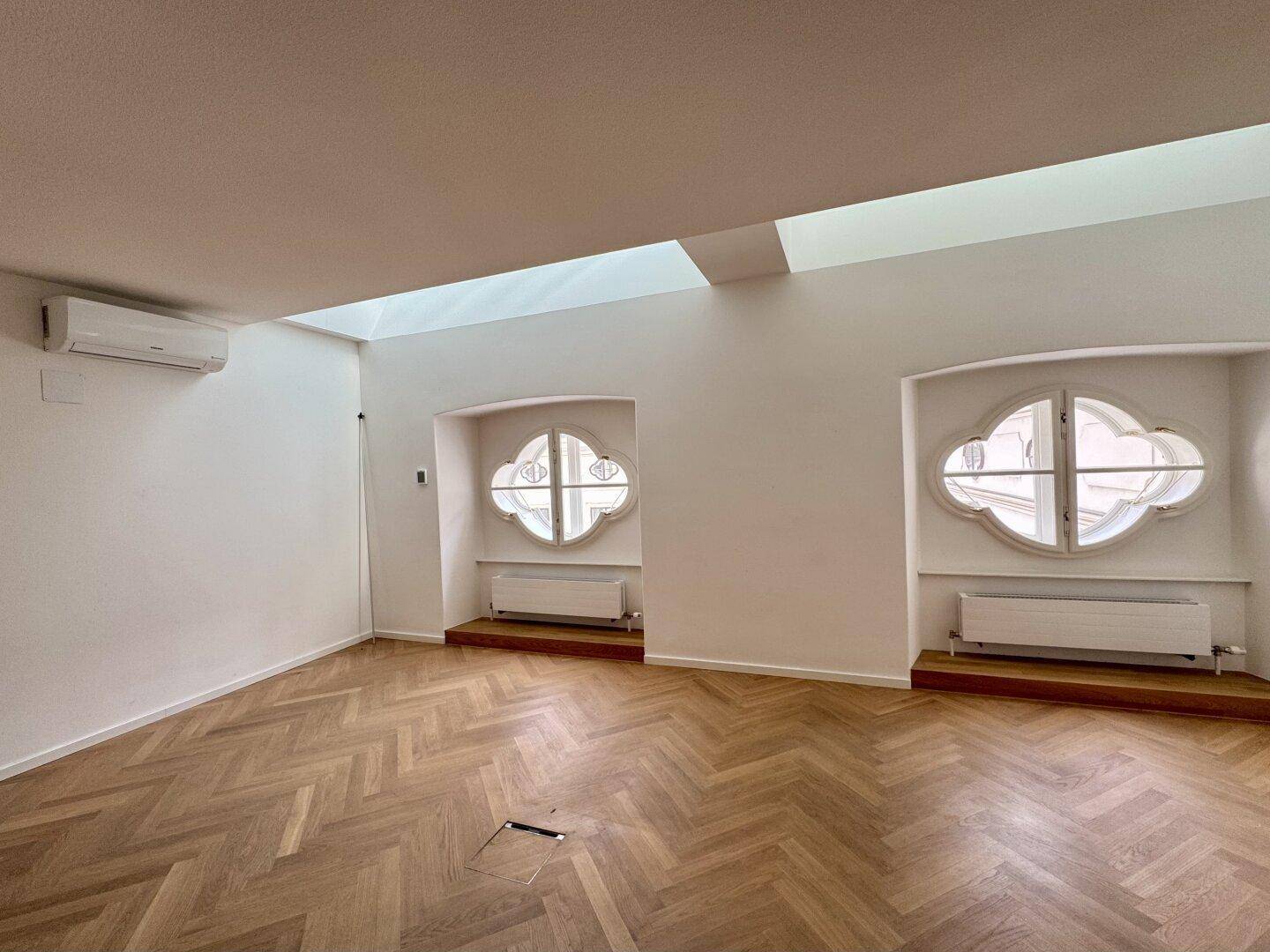 Heller Büroraum mit Fischgrätparkett, Klimaanlage und zwei dekorativen Rundfenstern.