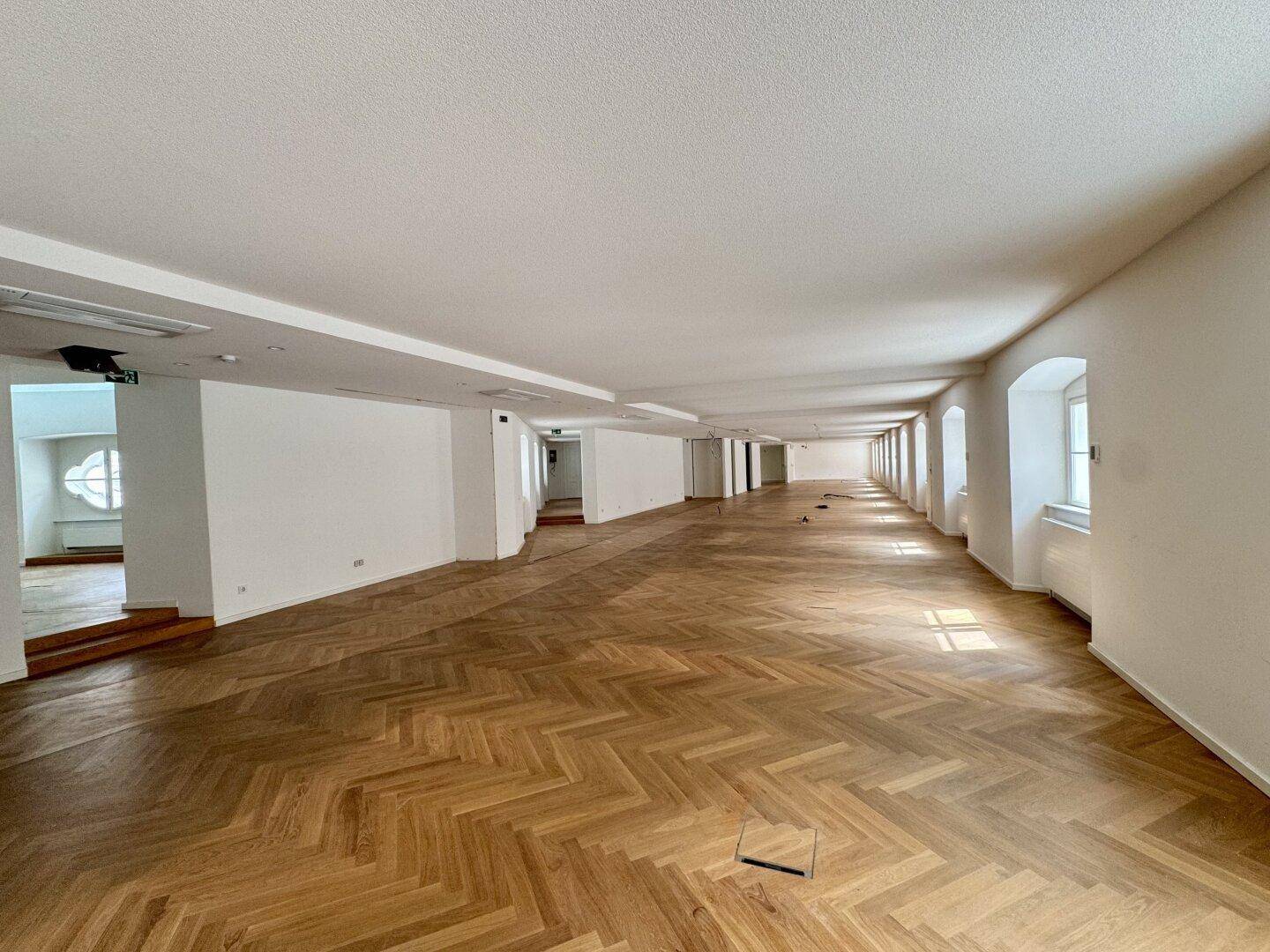Großer, leerer Büroraum mit Fischgrätparkett und langen Fensterreihen.