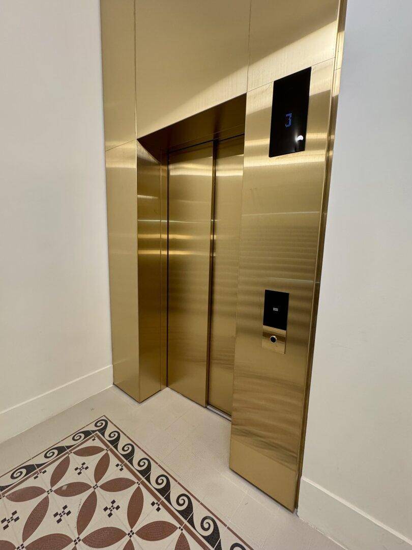 Moderner Aufzug mit goldfarbenen Türen und elegantem Fliesenboden im Eingangsbereich.