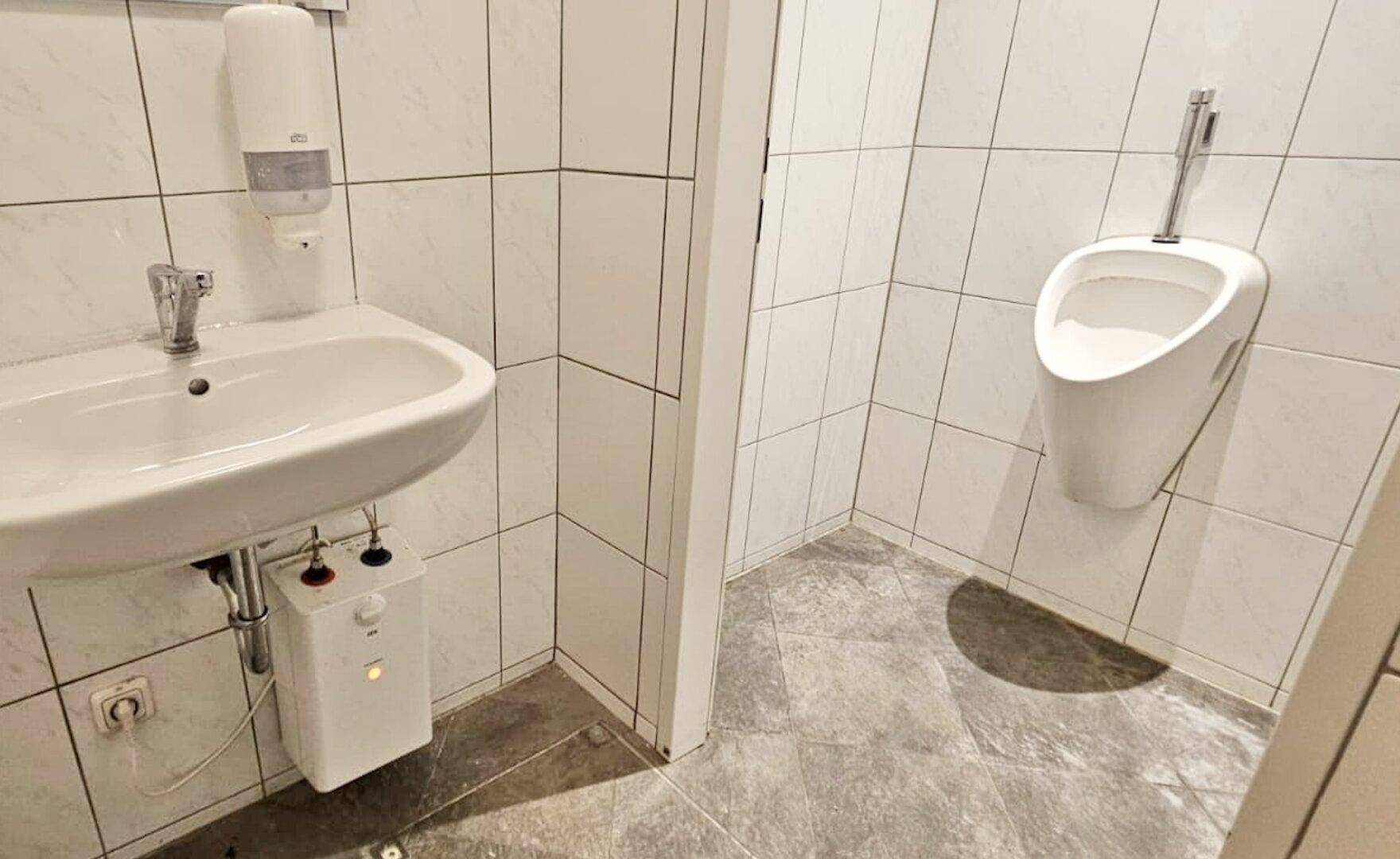 Kleine Toilette mit Urinal, Waschbecken und weißen Wandfliesen, funktional und sauber.