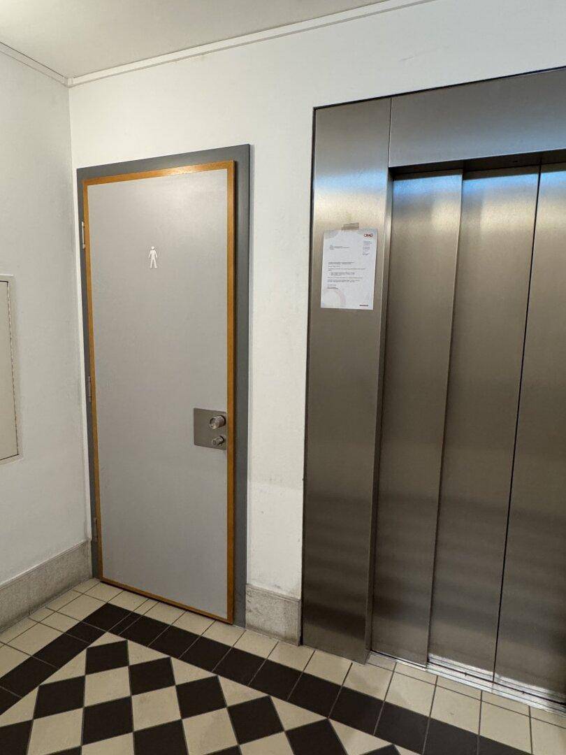 Flur mit modernem Aufzug und einer Tür, die zu einer Toilette führt, auf Schachbrettmuster-Fliesenboden.