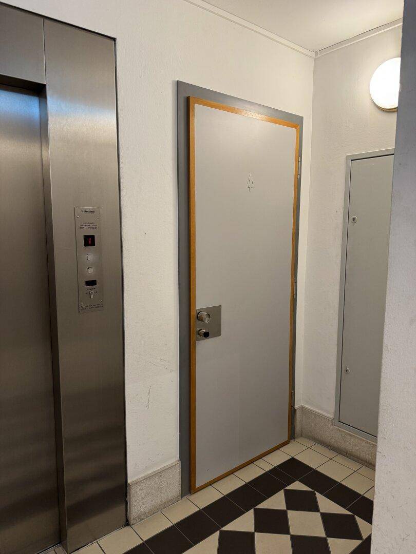 Moderner Aufzug neben einer Tür mit Piktogramm, auf einem Schachbrettmuster-Fliesenboden.