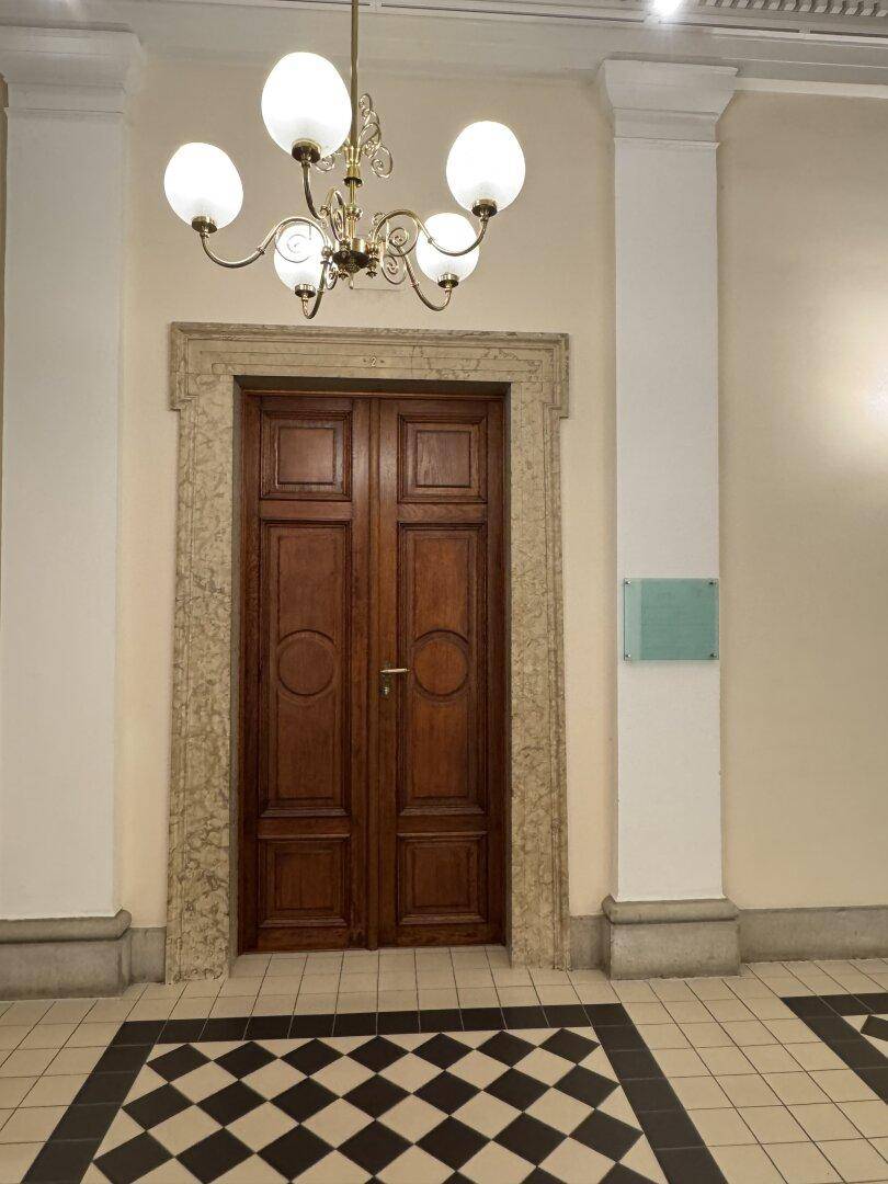 Detailansicht einer hohen Holztür mit Steinrahmen und Schachbrettmuster-Fliesenboden in einem historischen Flur.