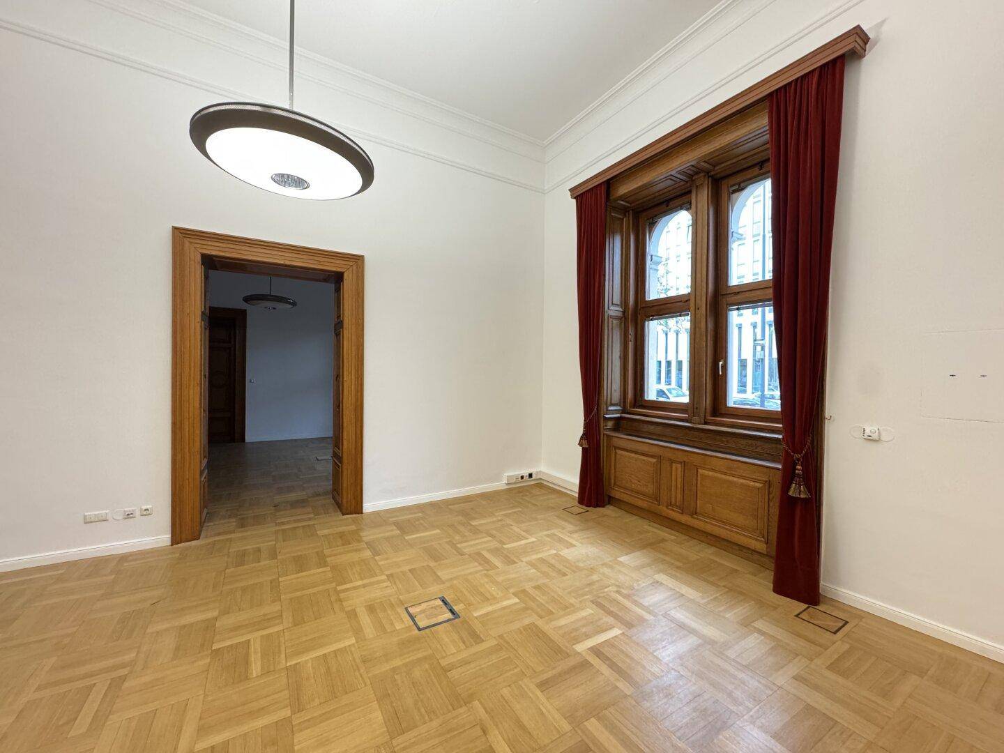 Heller Raum mit Parkettboden, hohen Fenstern und roten Vorhängen, ideal als Büro oder Arbeitszimmer.