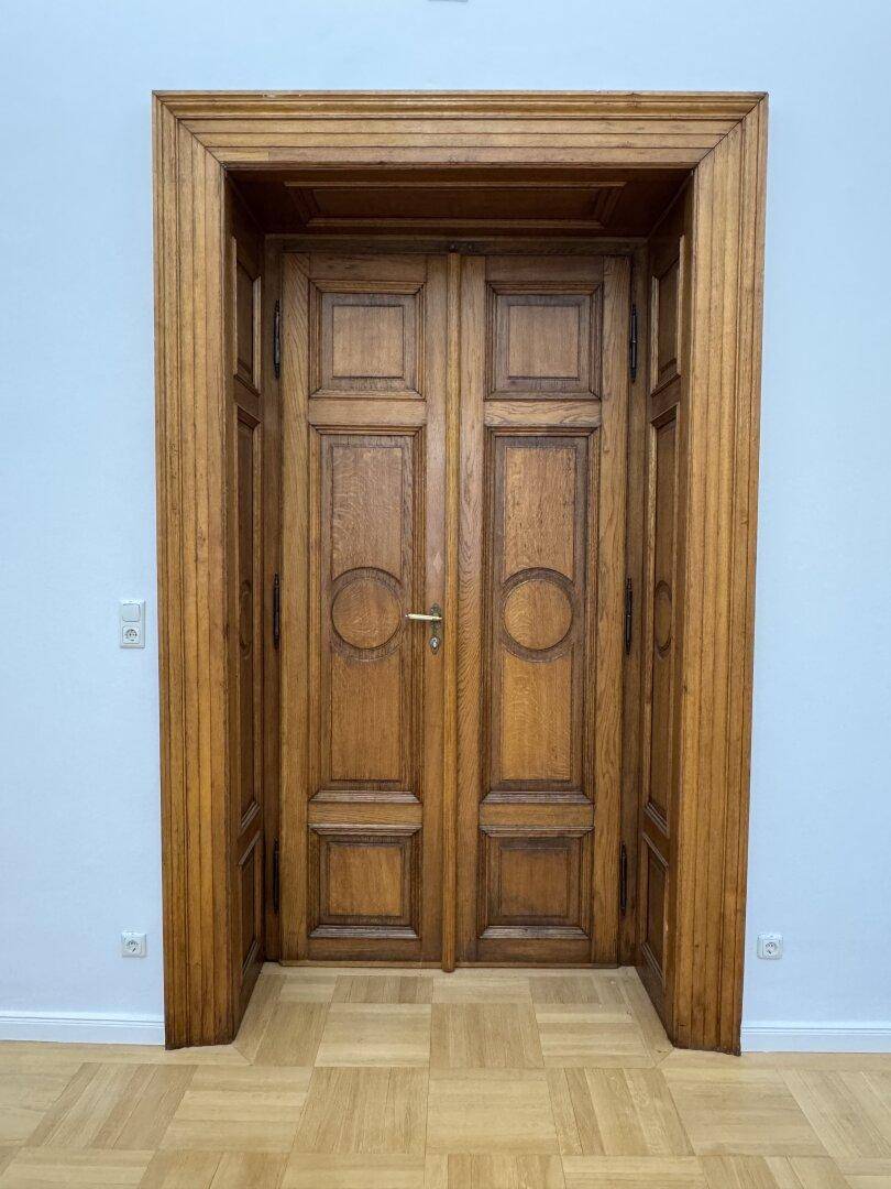 Eine detailreiche, hohe Holztür mit Zierrahmen und Parkettboden in einem historischen Gebäude.