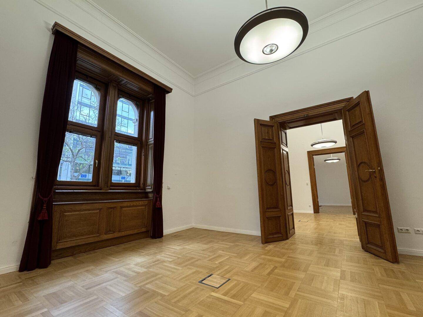 Geräumiger Raum mit Parkettboden, hohen Fenstern und doppelten Holztüren, ideal für Bürozwecke.