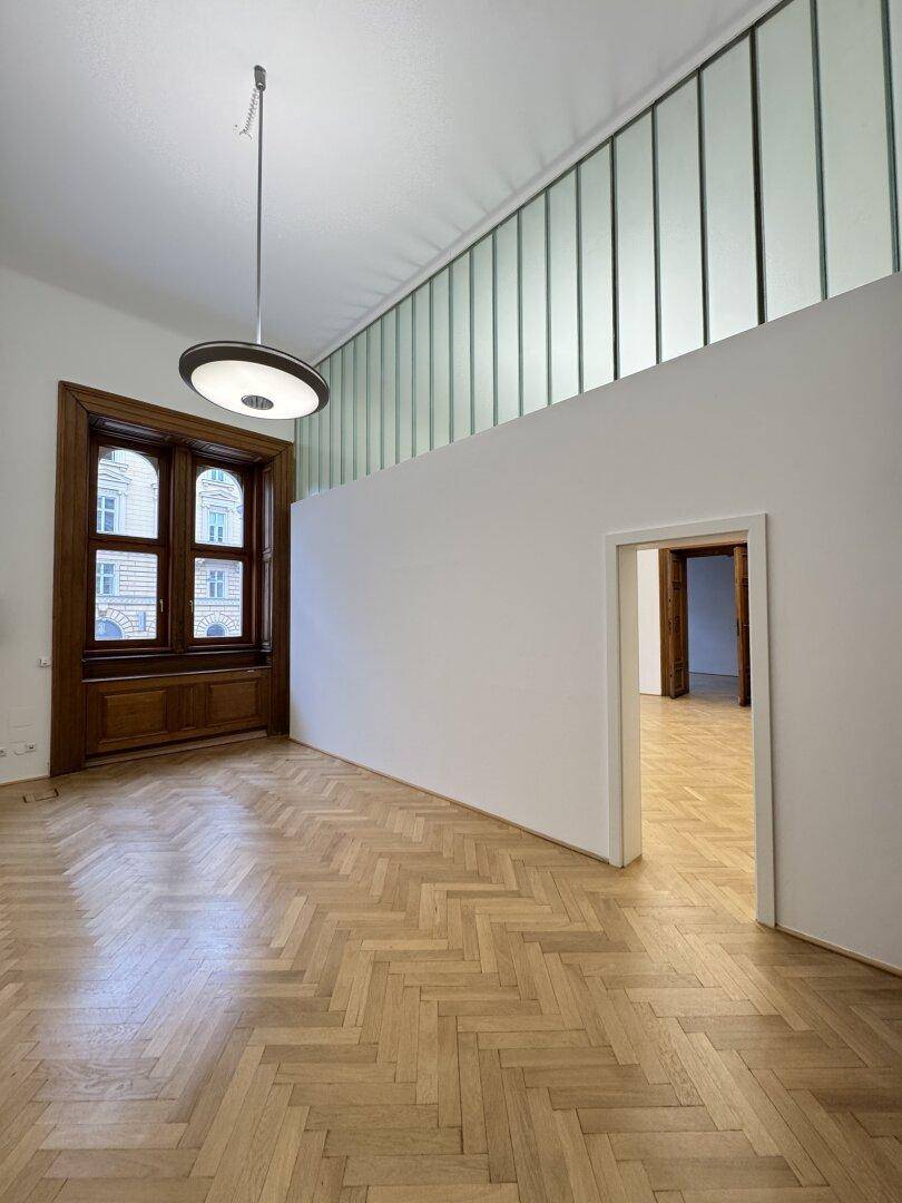 Heller Raum mit Fischgrätparkett, hohen Decken und einer oberen Glaswand für mehr Licht.