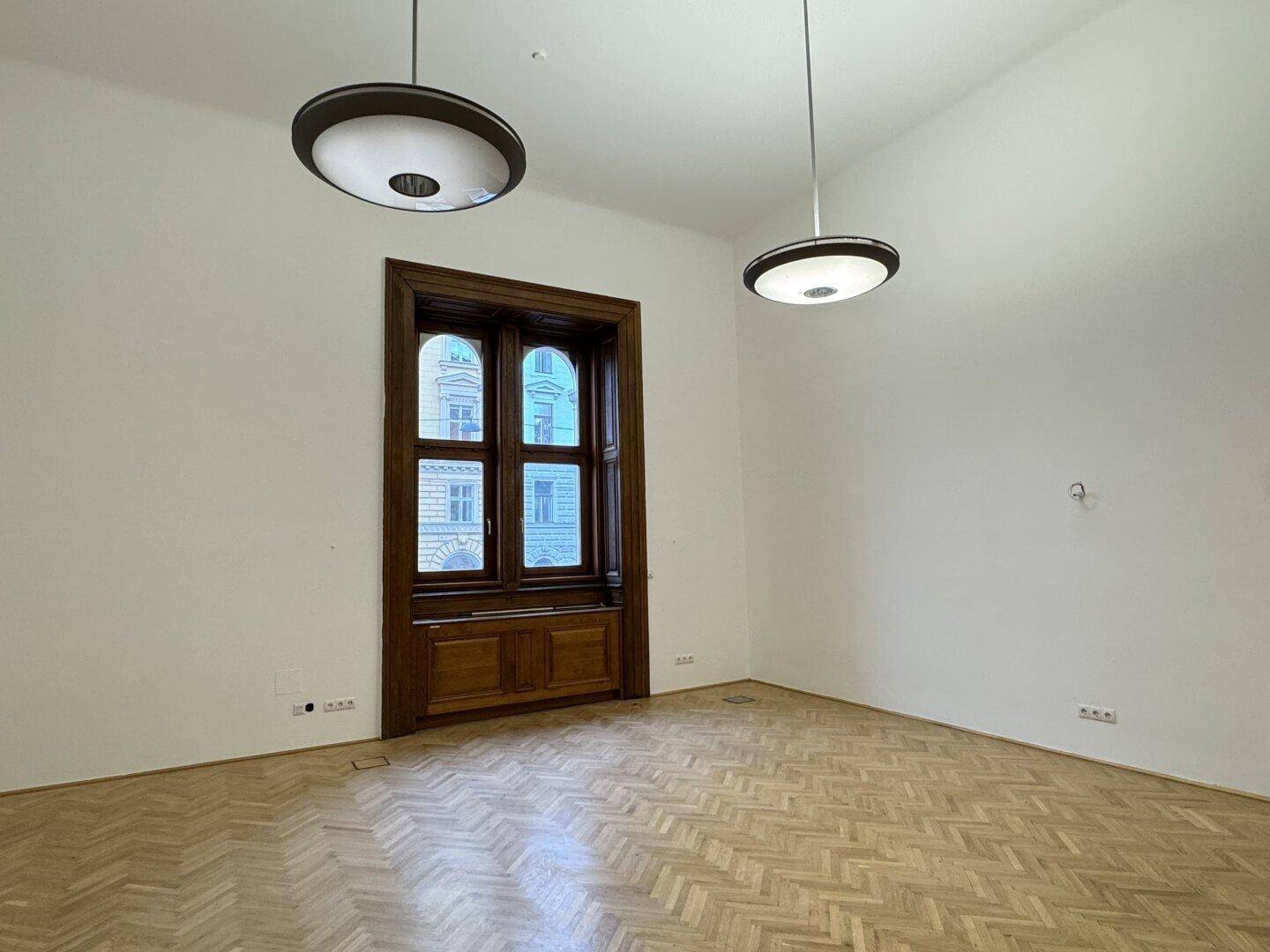 Heller Raum mit Fischgrätparkett, hohen Decken und einem großen Fenster mit Holzrahmen.