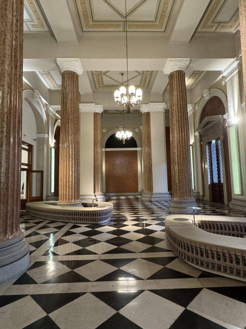 Prunkvolles Foyer mit hohen Säulen, Kassettendecke und einem auffälligen Schachbrettmuster auf dem Boden.