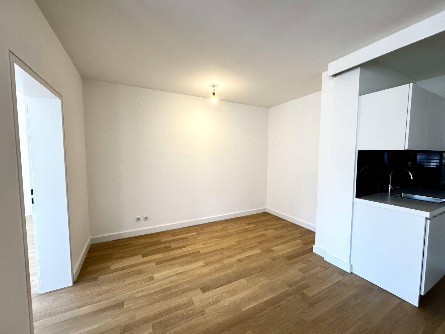 Heller Wohnbereich mit Parkettboden und weiß gestrichenen Wänden, angrenzend an eine kleine Küchenzeile.