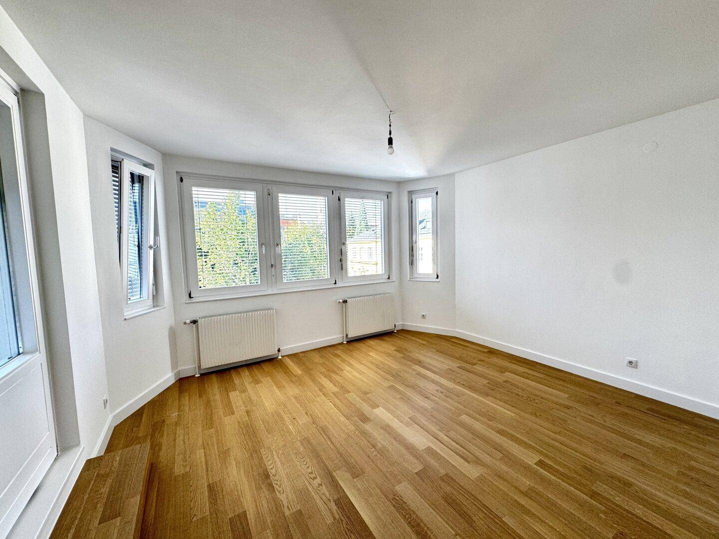 Geräumiges, leeres Wohnzimmer mit vielen Fenstern, Parkettboden und Heizkörpern unter den Fensterbänken.