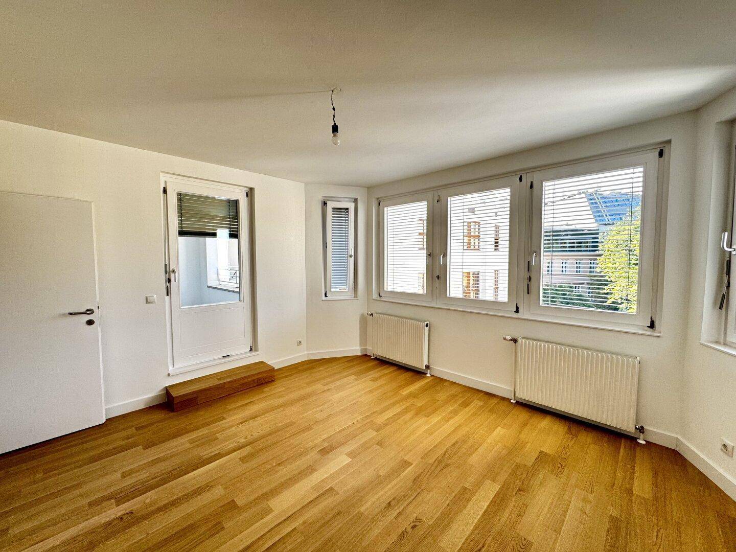 Lichtdurchfluteter Wohnraum mit Holzboden, großen Fenstern und einer Tür, die ins Freie führt.